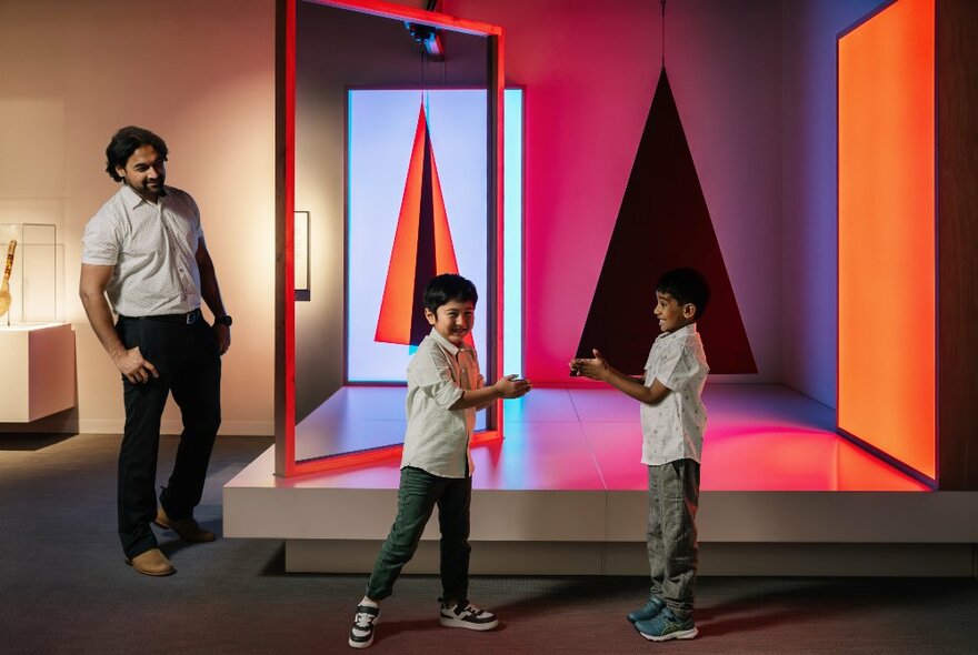 Two preschool aged children interacting with each other next to a low platform of mirrors and shapes, in a gallery space, while a smiling adult stands nearby.