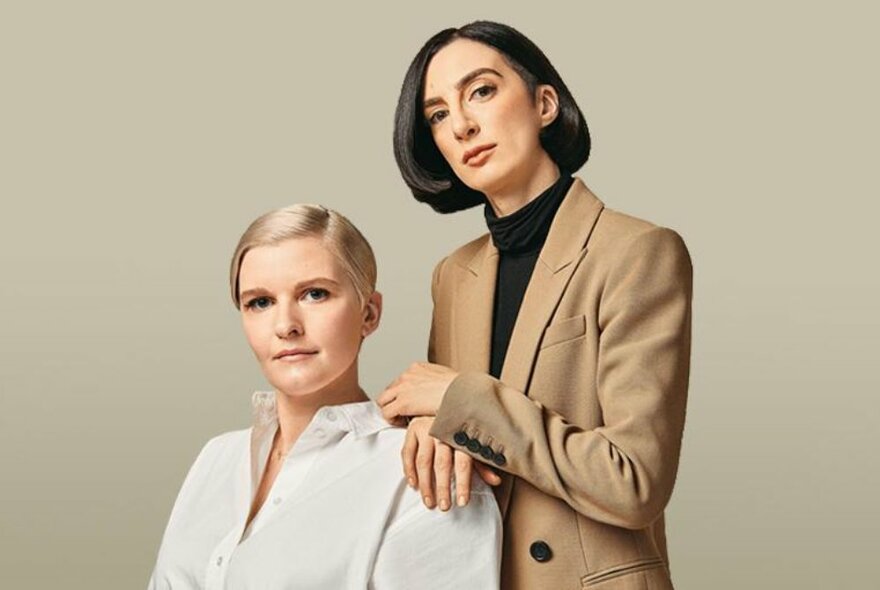 Friends and podcast hosts Chelsea Fairless and Lauren Garroni, one standing behind the other, in smart business like attire, against a pale neutral background.