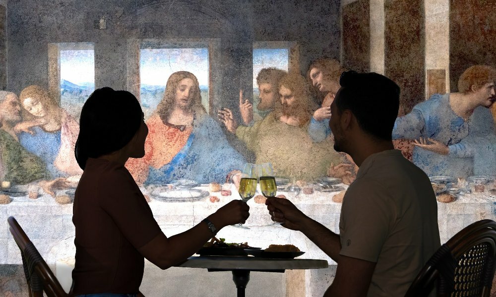 People clinking glasses at a small restaurant table with Da Vinci's last supper painting in the background.