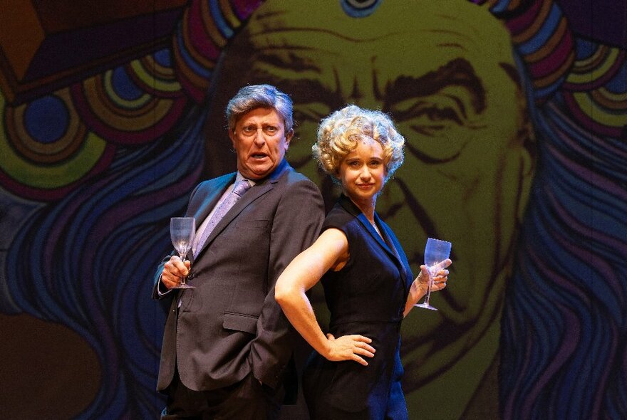 A male and female performer on stage, dressed in office attire, holding wine glasses and standing back to back.