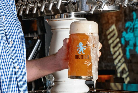 Close up of a bartender holding a freshly poured Bavarian beer which is spilling over the side of the large glass. 