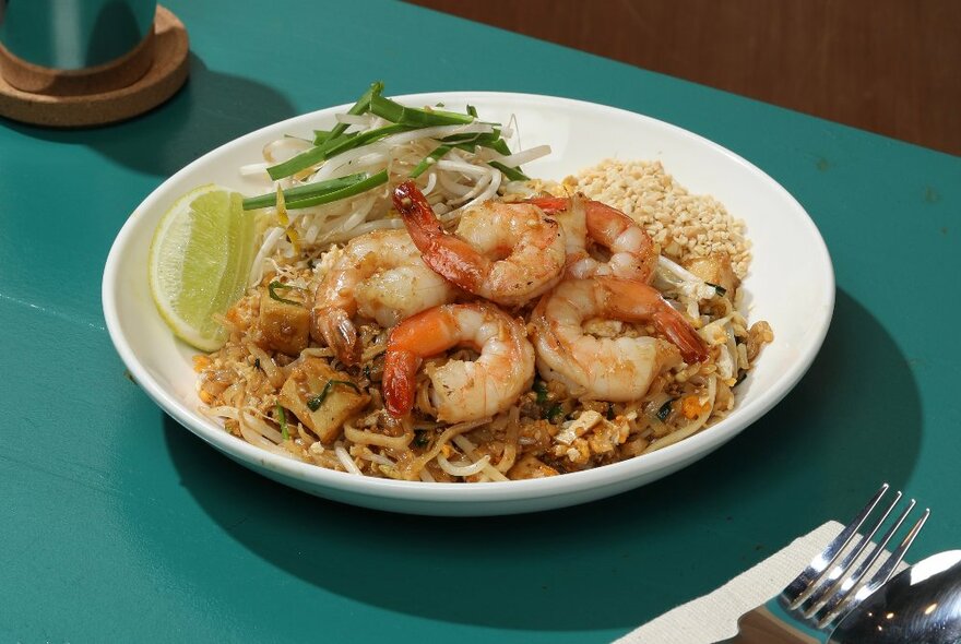 A dish of prawns and noodles in a low white bowl on a table, with cutlery.