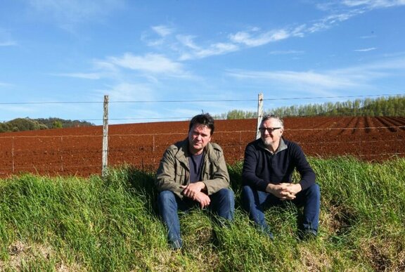 Two men, sitting on a grassy mound, in a farming field in Tasmania, with blue sky and rich red soil in the background.
