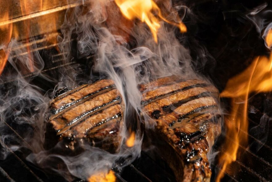 Two steaks sizzling over a char grill with smoke rising and flames visible.