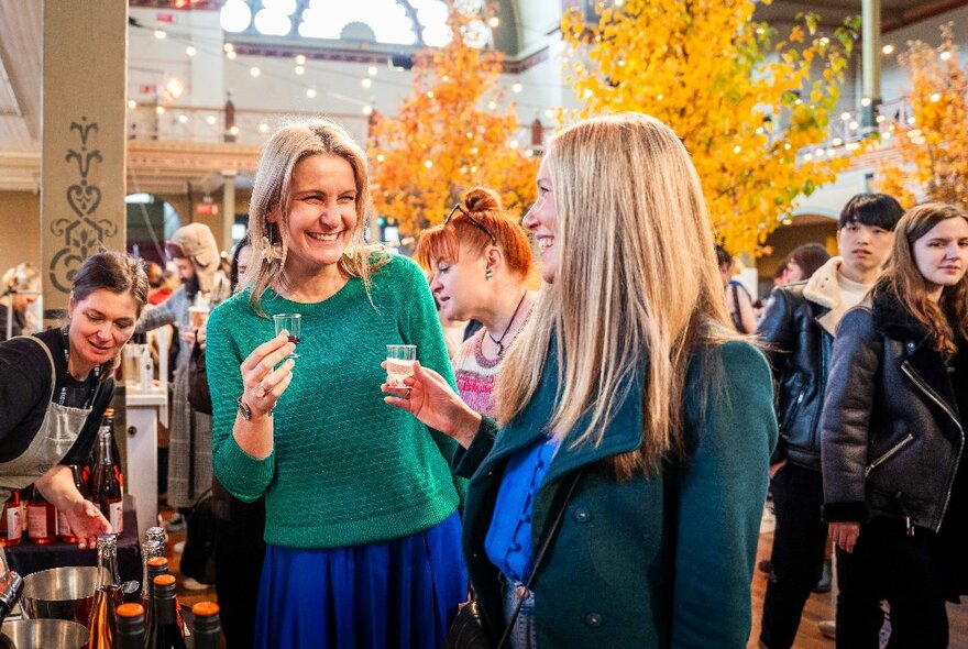 Two women at The Little Food Market at the Royal Exhibition Building, both holding small wine tasting glasses and smiling.