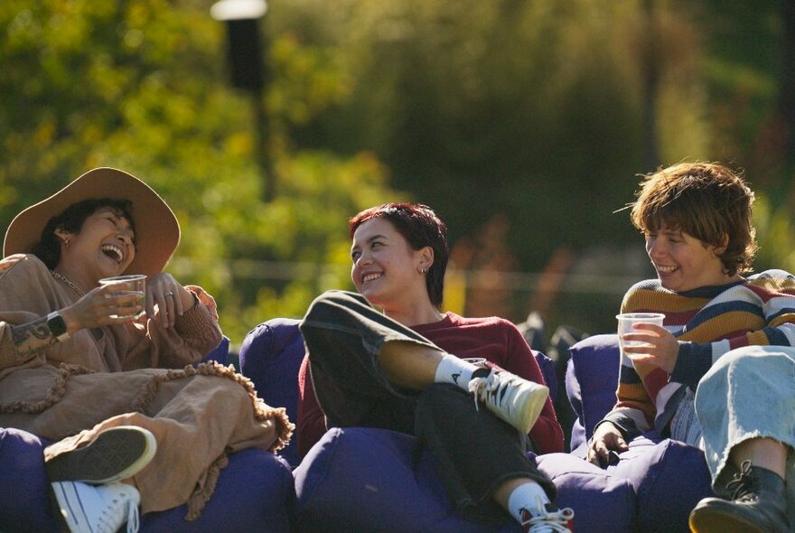 Three laughing friends sitting on bean bag couches with drinks in the Royal Botanic Gardens.
