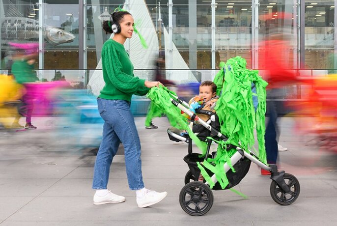 A woman wearing headphones pushes a pram that is decorated with fringed green paper as a baby pokes its head out.