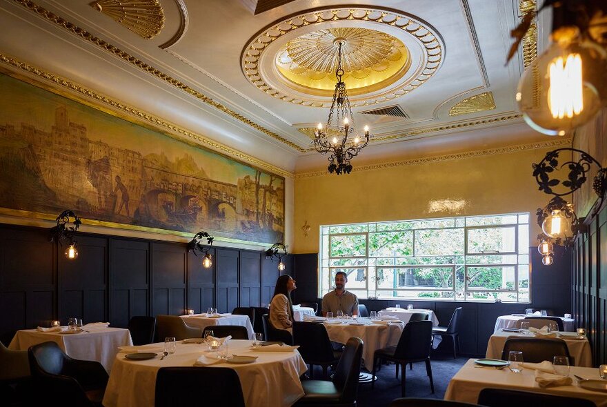 A couple dining in a sophisticated restaurant with a large mural and chandeliers.