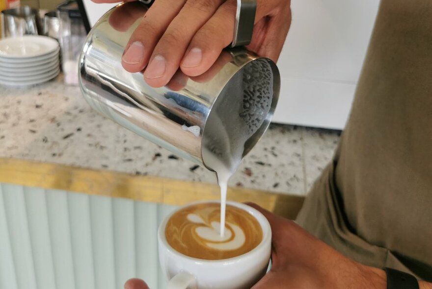 Hand pouring heated milk from a metal jug into a cup of coffee on a cafe counter.