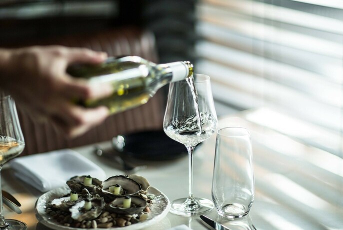 A hand pouring a glass of white wine beside a plate of oysters.