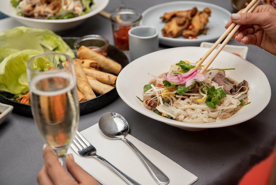Someone lifting noodles with chopsticks at a table with multiple plates of Vietnamese food, champagne and condiments. 