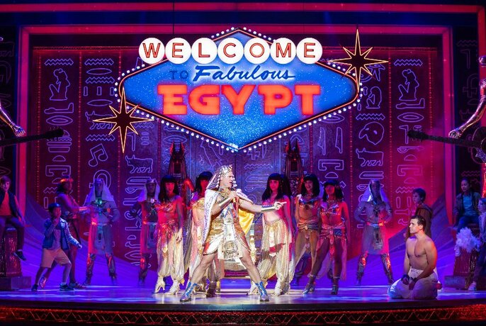 Cast of performers in costume posed on a theatre stage, under a neon sign that displays the words 'WELCOME TO FABULOUS EGYPT'.