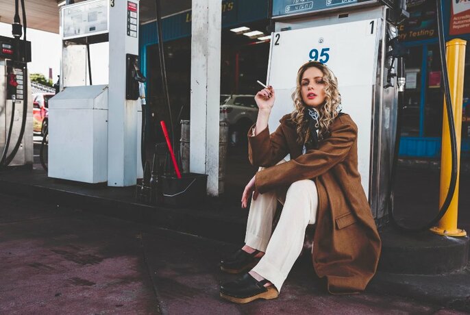 Model wearing a brown coat and smoking a cigarette next to a petrol bowser in a service station.