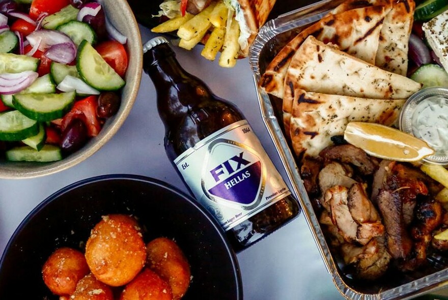 A bottle of beer surrounded by Greek salad, donuts, pitta and meat.