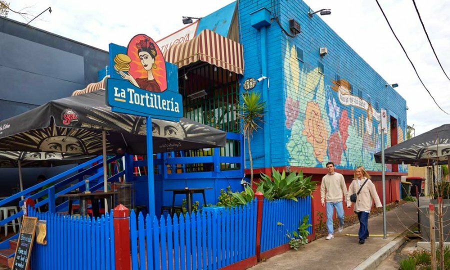 Couple walking side by side passed blue picket fence and blue brick building with the 'La Tortilleria' sign.