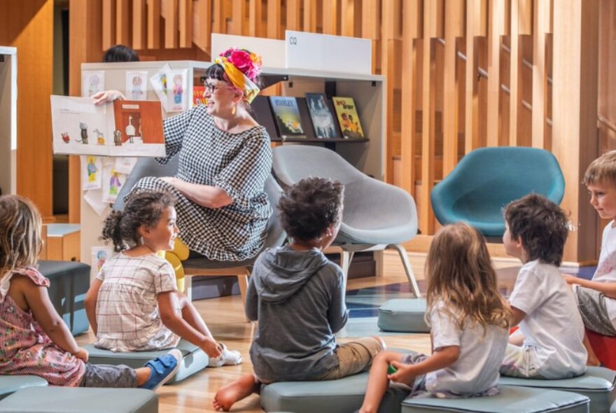 A person seated on a chair, reading from an open picture book they are holding up to a group of preschool-aged children seated on floor cushions listening and watching.