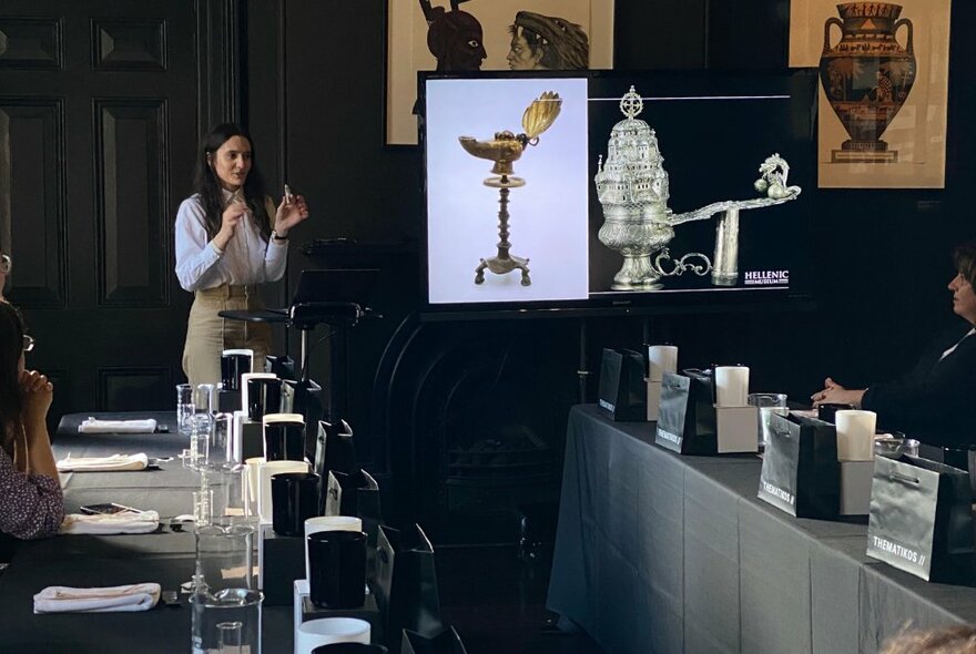 A woman presenting with visuals in front of a small group of people at long tables.