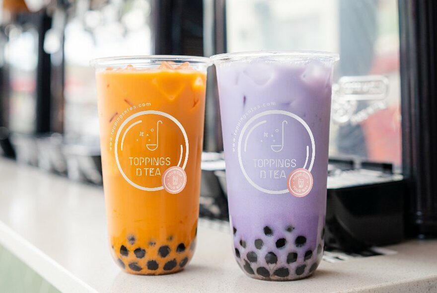 One orange and one lavender bubble tea waiting on a counter. 