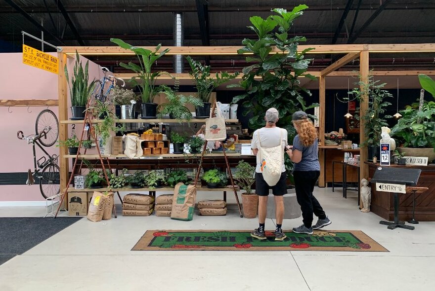 Two people browsing wooden shelves in warehouse-style cafe/pantry interior, with many green plants.