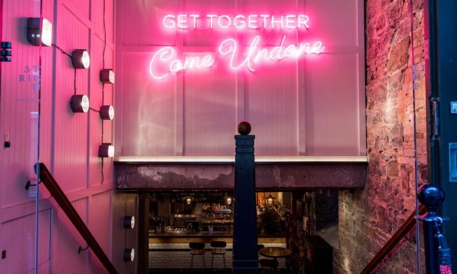 A flight of stairs leading to a basement bar with a neon sign reading 'get together come undone'.