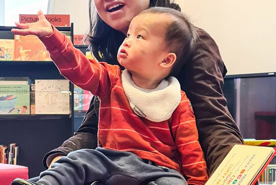 Young toddler seated on an adult's lap, holding  their arm up in the air, in a library setting with a shelf of picture books behind them.