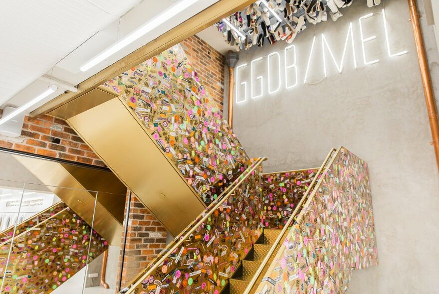 A golden stairway adorned in colourful stickers to look like confetti, with a neon sign on the wall reading GGDB/MEL