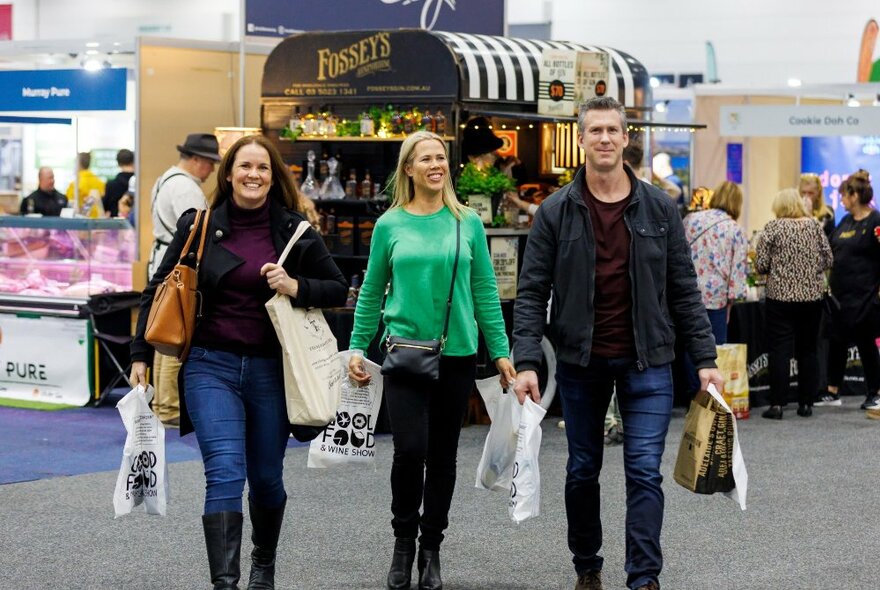 Three expo-goers carrying bags, standing in an event space with food stalls.