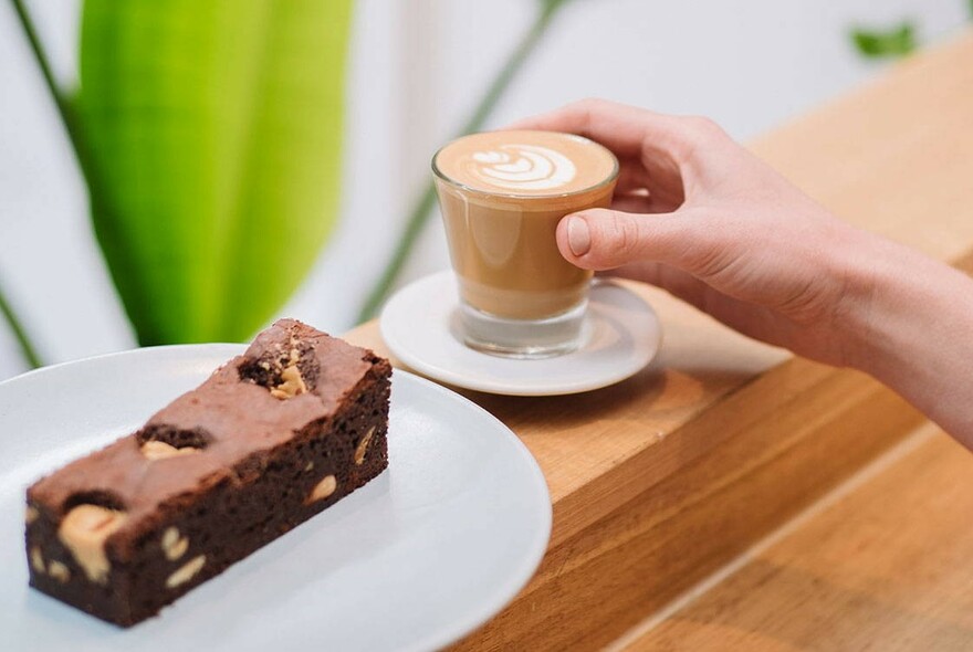 Hand holding a caffe latte on a saucer next to a chocolate brownie on a plate, on a wooden counter.