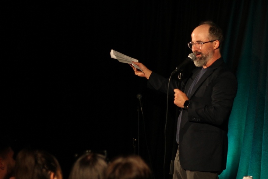Middle-aged balding man in a suit, holding a notebook in his hand, standing on a stage and talking into a microphone.