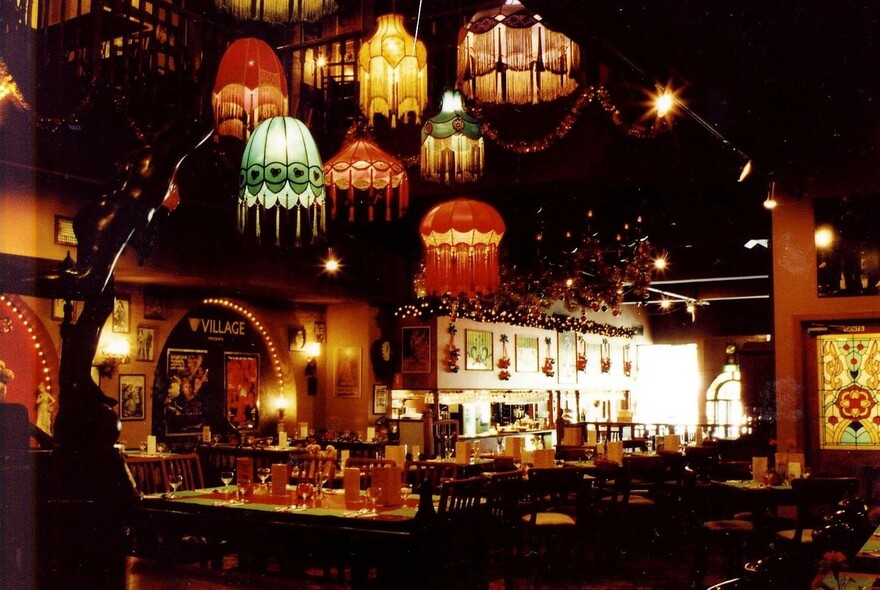 Tasselled lampshades hanging from the ceiling of a moodily lit restaurant, with dark tables and chairs.