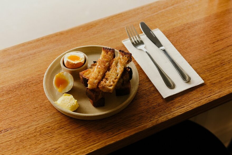 Plate of soft-boiled dippy eggs, toast rectangles, and a knife and fork resting on a white napkin, on a timber table top.