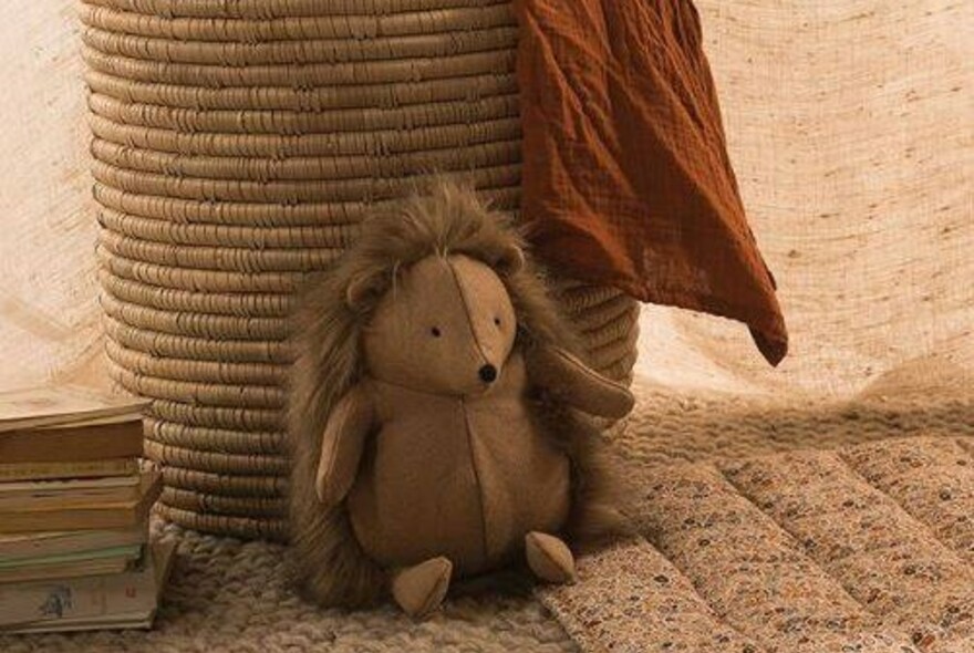 Toy porcupine resting against a basket, with floral eiderdown to the right and a small stack of books to the left.