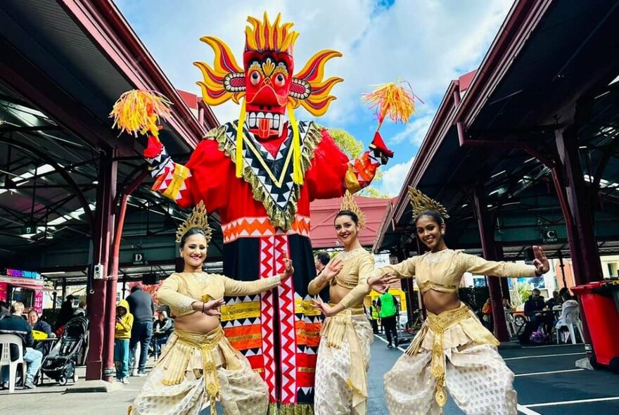 Sri Lankan dancers performing in the Queen Vic Market sheds next to a tall dance puppet figure.