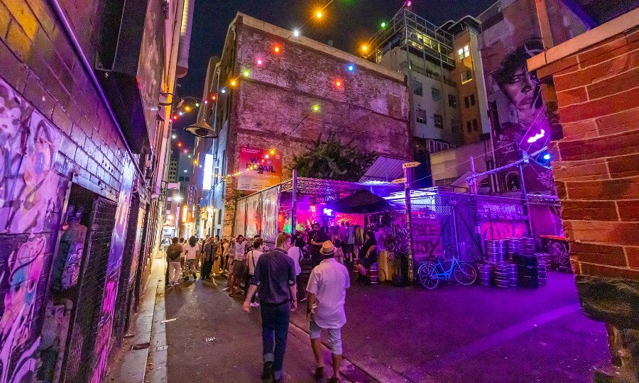 People walking down a laneway at night towards a busy laneway bar with colourful lights.
