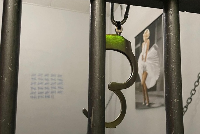 Handcuffs hanging from prison cell bars, date count down marks and Marilyn poster on walls behind.