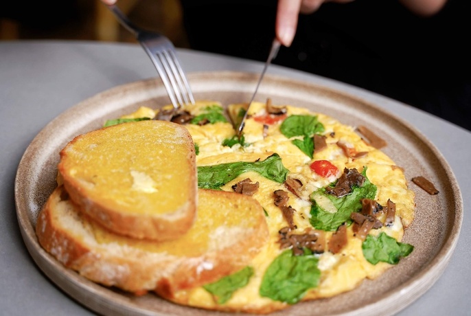 Knife and fork cutting into omelette on light brown ceramic plate, with two slices of buttered toast to side.