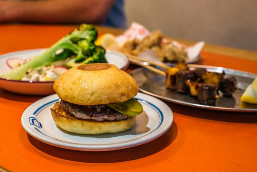 Close up shot of burger on an orange table next to chicken skewers and vegetables.