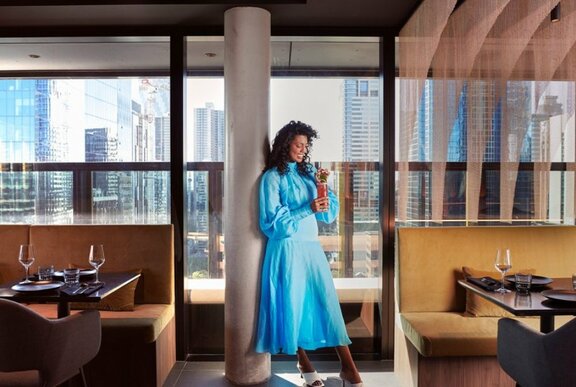 Woman wearing a blue dress holding a drink with a straw in a hotel cafe with banquette seating and tables.