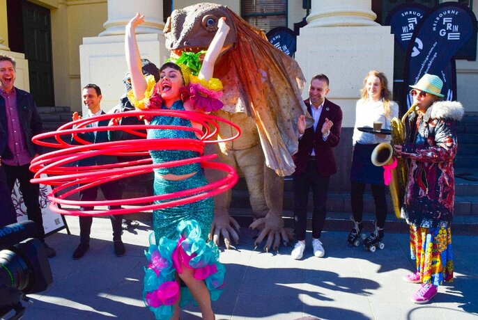 Person in aqua frock hula-hooping with multiple bright pink hoops, large lizard puppet behind, sax player to right.
