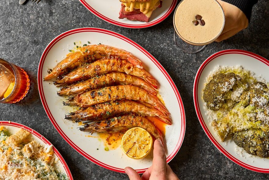 A hand positioning a halved lemon on a plate of cooked prawns next to other dishes.