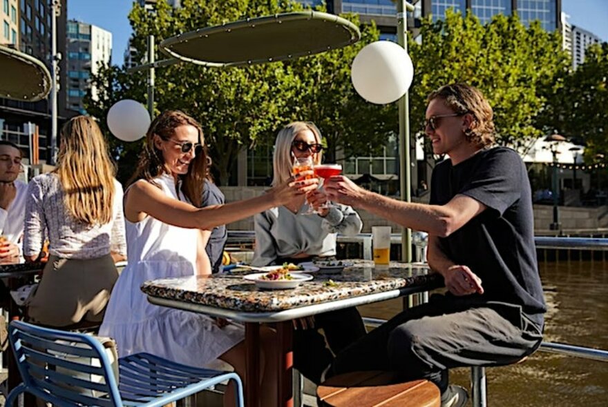 People seated at an outdoor table by the Yarra clinking glasses.
