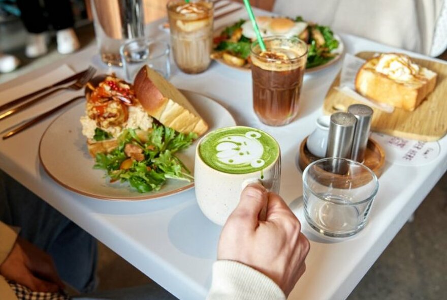 Side shot of table with scrambled eggs, spinach and thick toast, iced coffee, thick buttered toast. male hand holding mug of matcha with bear latte art.