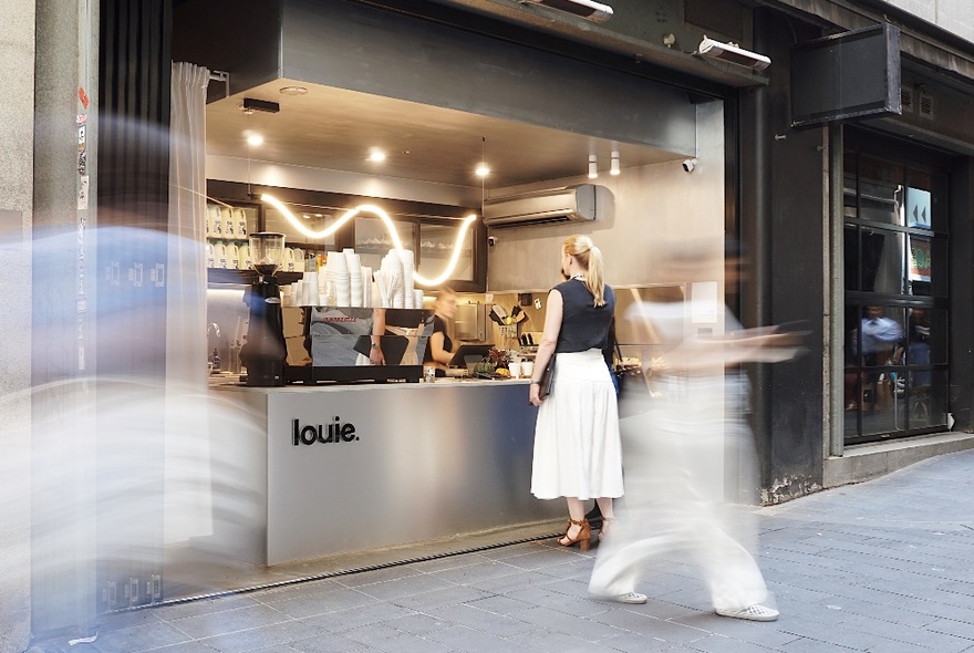 A blurred motion picture showing a woman in a long white skirt and black sleeveless top ordering at a small cafe counter. 
