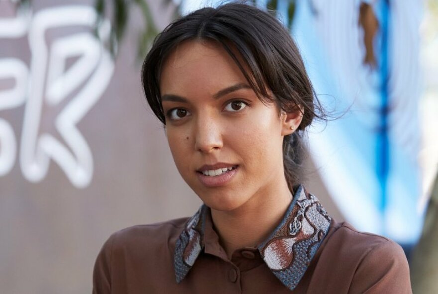Model looking at viewer, slightly smiling, wearing a brown blouse with an embroidered collar.