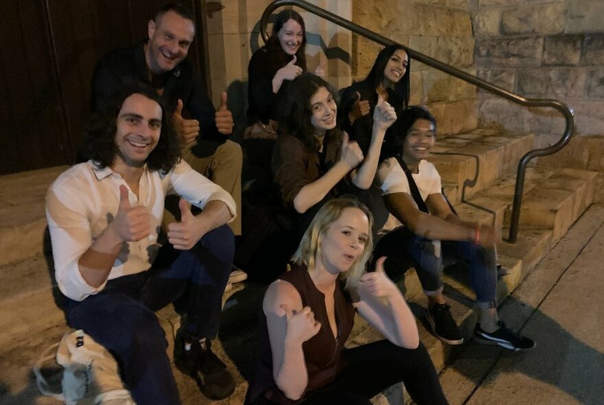 A group of people sitting on stone steps at night with thumbs up. 