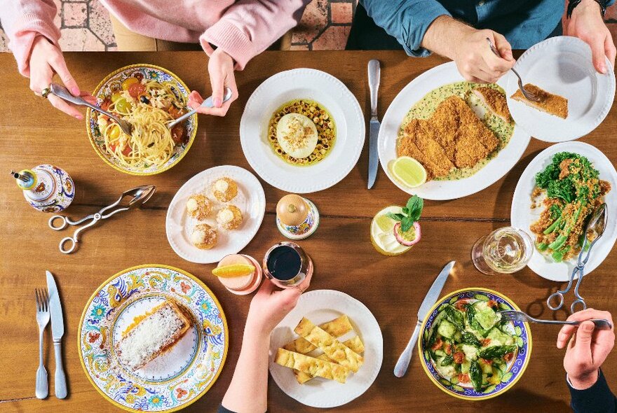Overhead view of assorted plates of Italian food on a table with people using cutlery and holding glasses of wine.