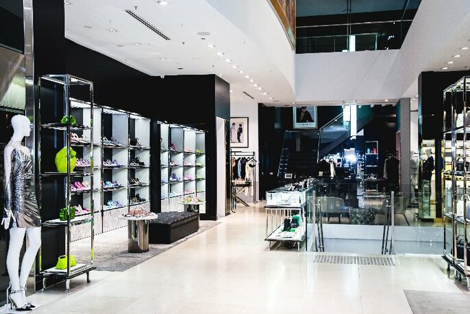 Interior of Harrolds store, a large open space, white walls, white ceiling and tiled floor, with racks of glass shelves to the left displaying leather bags.