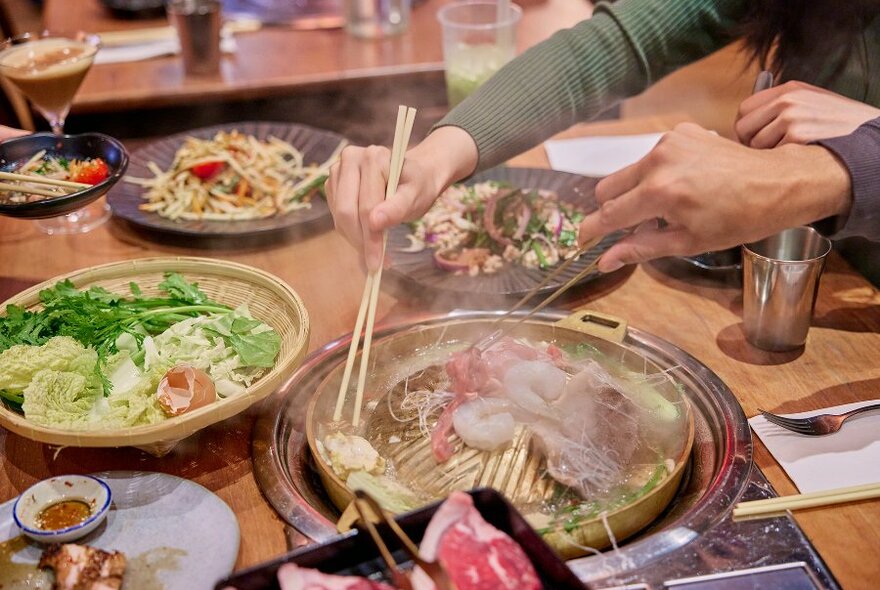 People reaching for meat on a gold mookata grill surrounded by Thai dishes.