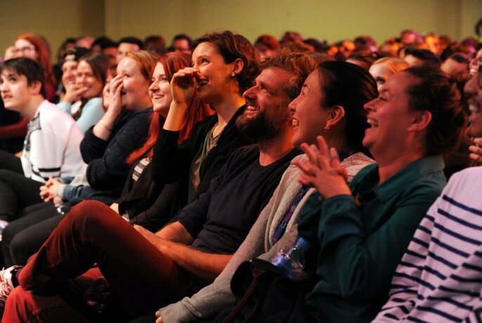 Audience seated in rows, laughing and smiling and looking towards the stage.
