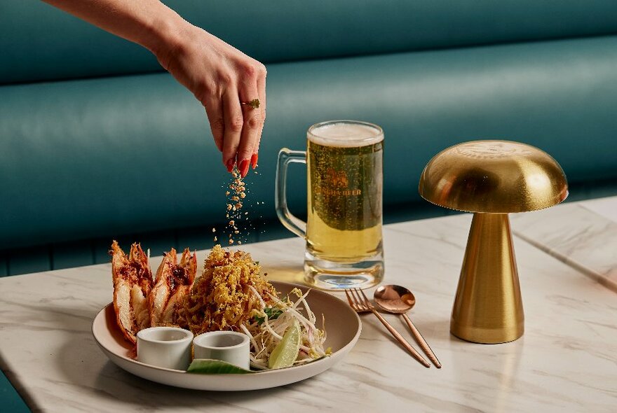 A hand sprinkling on garnish on a dish of prawns  with a pint of beer nearby and a small gold mushroom table lamp. 